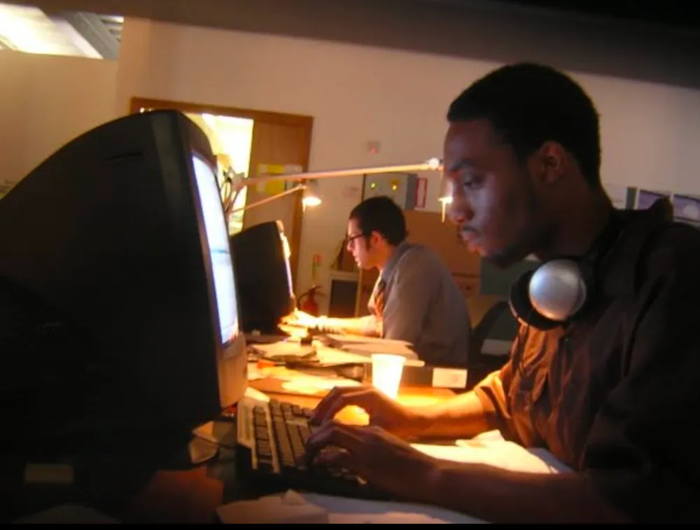 Two people working at computers in a dimly lit office; one has headphones around his neck and is typing.