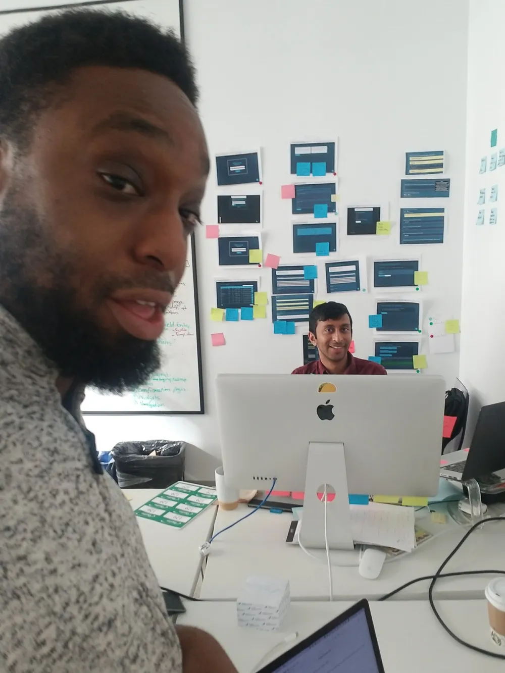 A man in a gray sweater turns toward the camera in the foreground. Behind him a colleague smiles from behind a desktop monitor, in front of a wall of printed screen mockups with sticky notes.