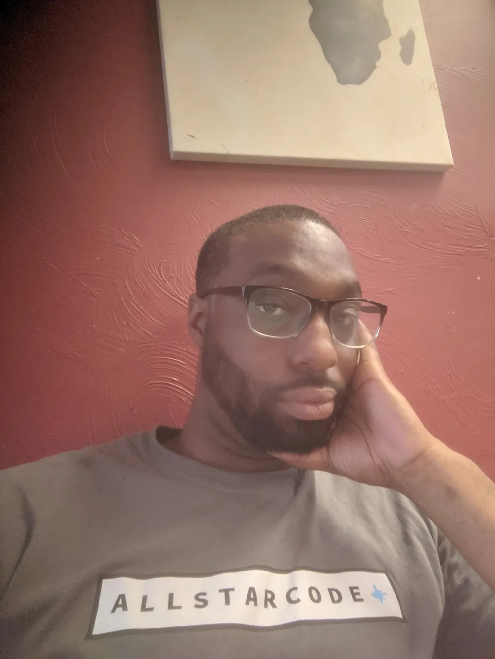 A man with glasses and a beard rests his cheek on his hand, wearing a gray ALLSTARCODE t-shirt in front of a red textured wall.