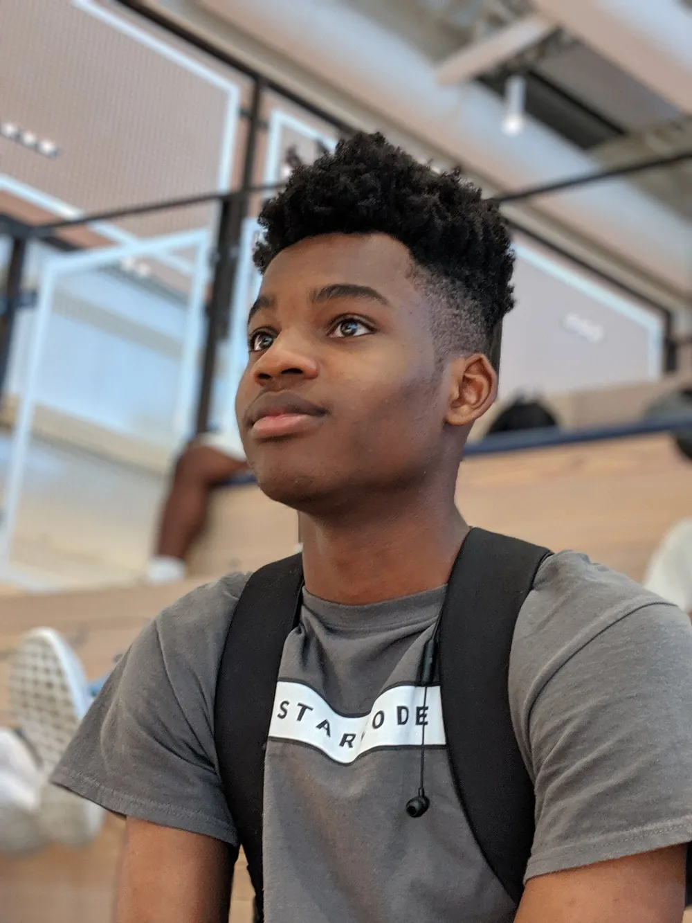 A young person in a gray t-shirt with a backpack, photographed from below as they look off to the side, seated on wooden bleacher steps.