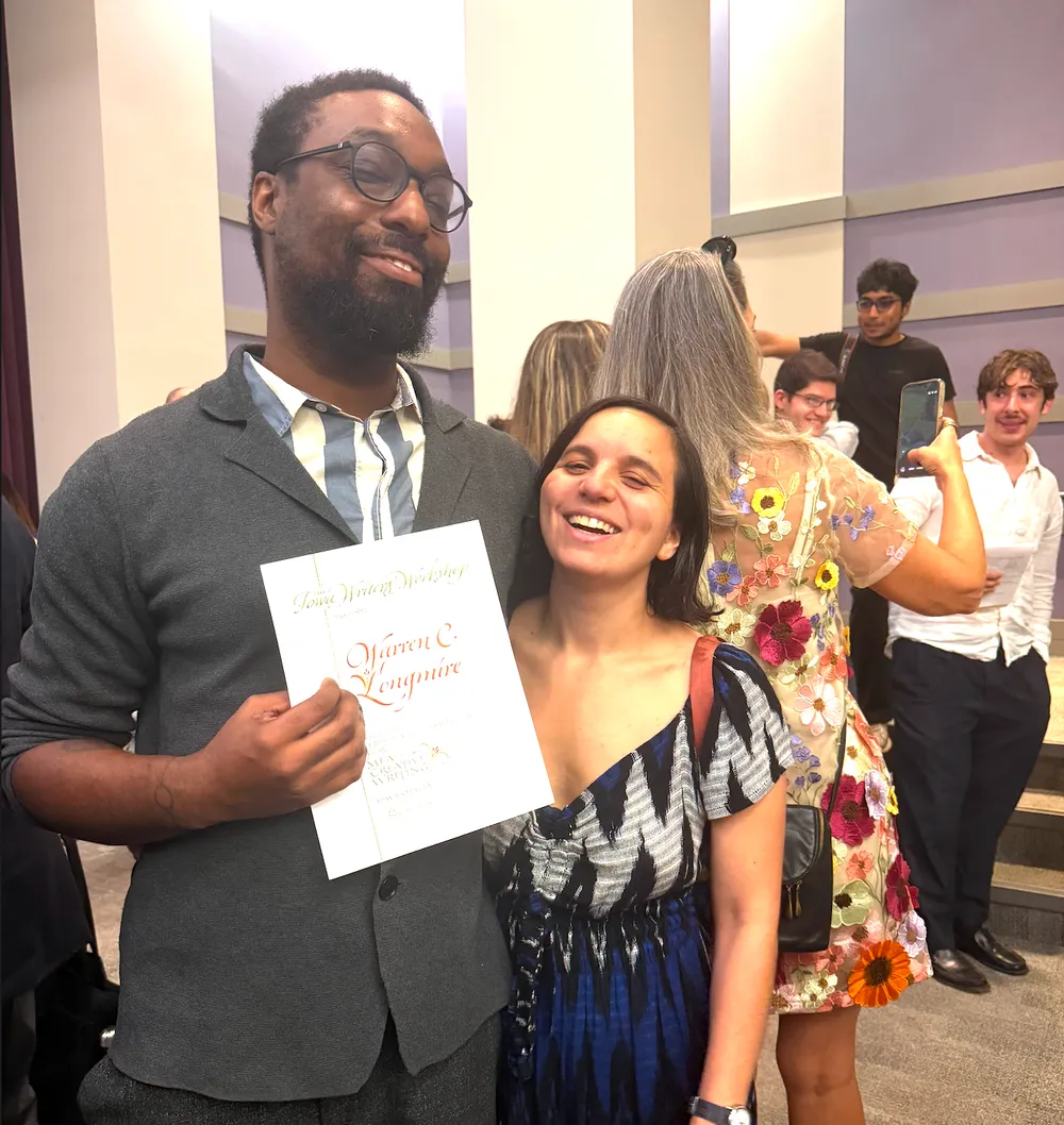 A smiling man in glasses and a gray cardigan holds up his diploma from the Iowa Writers' Workshop while a laughing woman leans against him, with other graduates and families behind them.