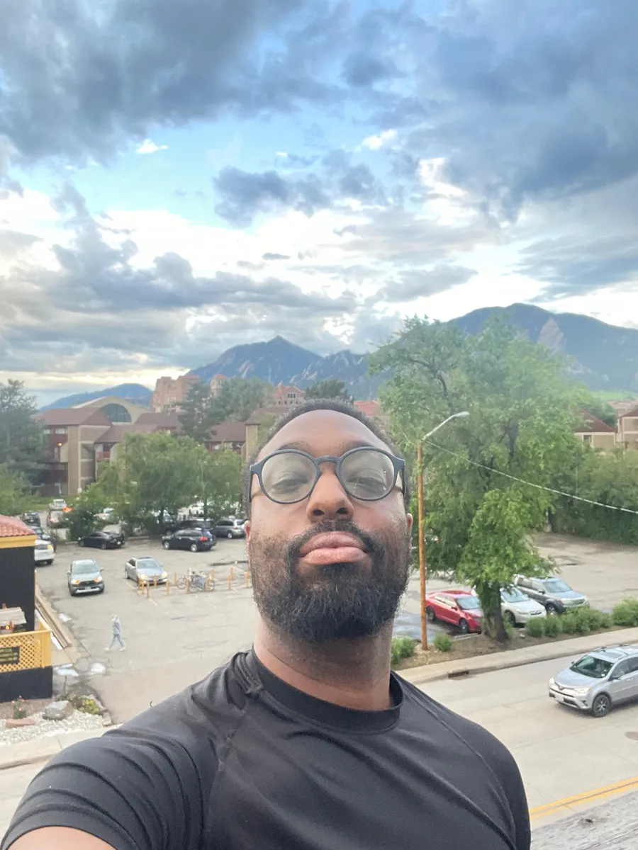 Warren C. Longmire, in round glasses and a black shirt, with cloudy skies and mountains behind him